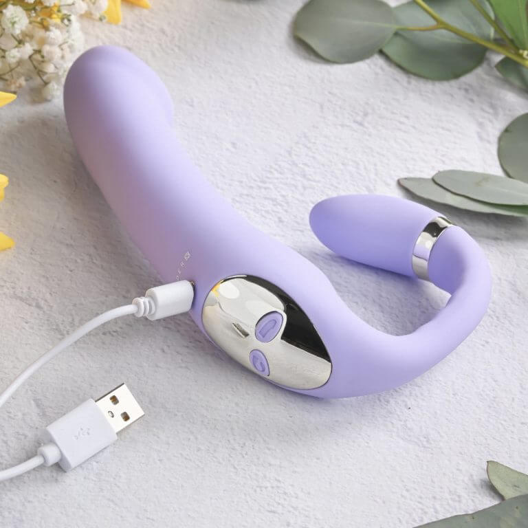 The Gender X Orgasmic Orchid lays flat on a texture surface with greenery around. The vibrator is plugged into its magnetic charging cable. The USB end of the other side of the charging cable is also shown in frame. | Kinkly Shop