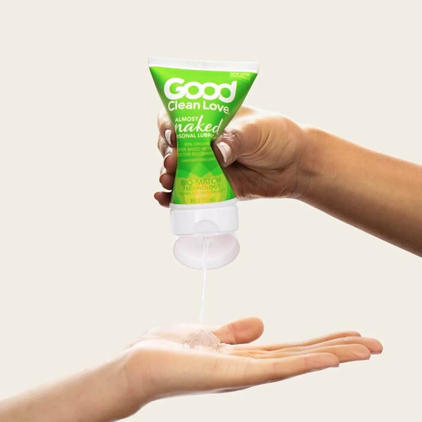 A hand holds the Good Clean Love Almost Naked Organic Lubricant - 4OZ and squeezes the bottle. Another hand is held open underneath the bottle to have a pile of lube squirted into their hands. The lube looks somewhat gel-like and makes a small pyramid in the hand. | Kinkly Shop