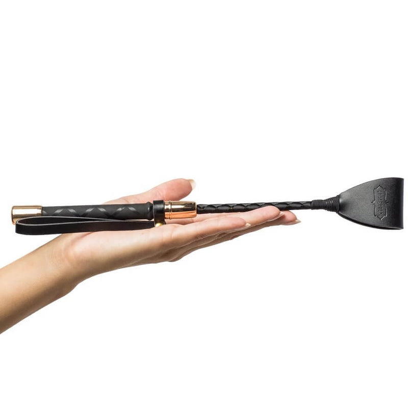 A hand, laying flat, displays the included riding crop. The crop is much longer than their palm, and it probably is about the length of a forearm. It has the Kama Sutra logo emblazoned on the riding crop tip, and it is a black/gold color scheme. It is much shorter than the standard riding crop. | Kinkly Shop
