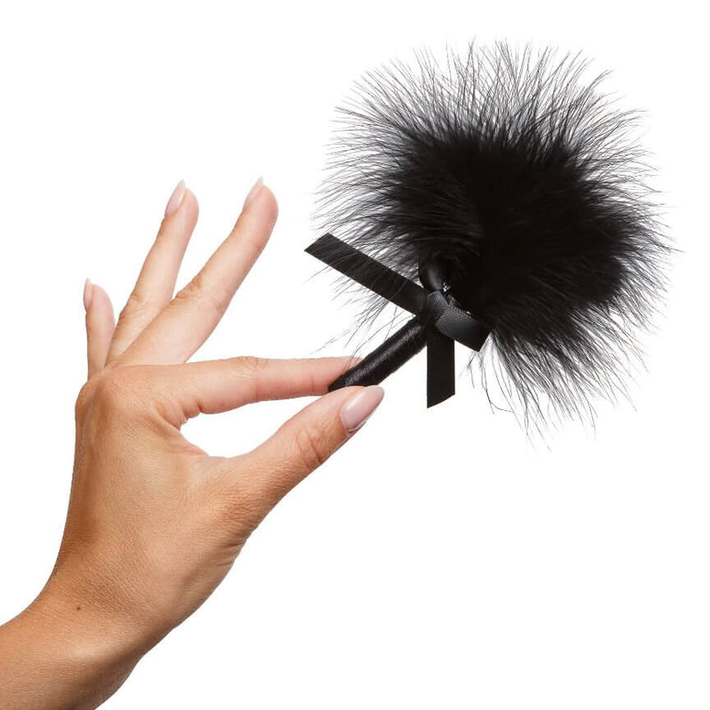 A hand holds the included feather teaser in the Kama Sutra Getaway Kit. It is about the length of a hand and includes a small black bow. | Kinkly Shop