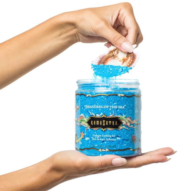A hand is flat and holding the container of the Kama Sutra Treasures of the Sea Bath Salts on it. The other hand is scooping out some of the bath salts with the included seashell scoop. | Kinkly Shop