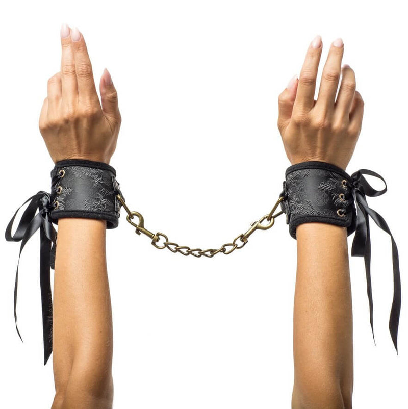 Two hands wear the cuffs included in the Kama Sutra Trust Me Erotic Playset. The cuffs are fastened with ribbons and have a satin-like finish. A gold chain with clips on both sides connects the cuffs to one another. | Kinkly Shop