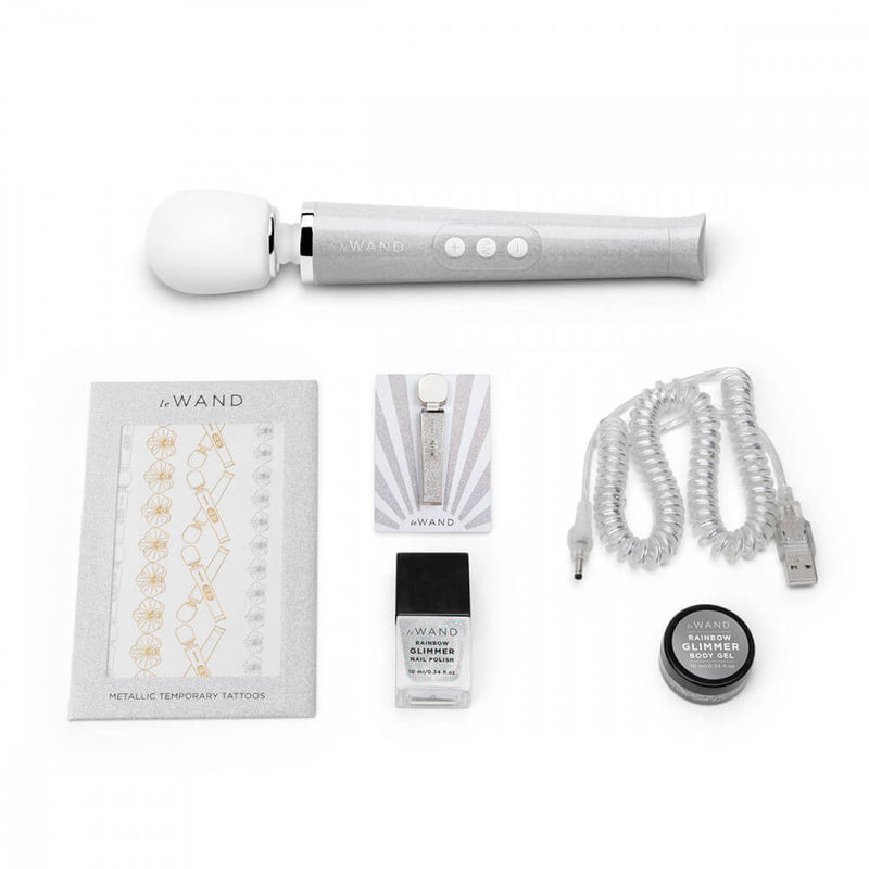 The White Le Wand All That Glimmers Wand Massager laid out next to everything that comes with the petite wand massager. That includes the color-coded charging cable, a glittery body gel, a matching enamel pin, a glittery nail polish, and a set of color-matching metallic temporary tattoos. | Kinkly Shop