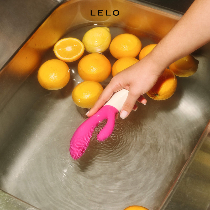 A hand holds the LELO INA Wave 2 Cerise inside of a sink full of water and oranges. The rabbit vibrator is currently on and vibrating which is making large waves inside of the water. | Kinkly Shop