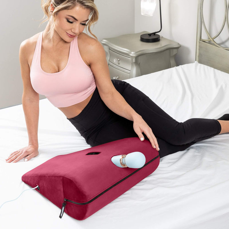 A person sits on their hip on a bed. The Liberator Axis Magic Wand Toy Mount is sitting in front of them with a wand massager tucked into the shape. The cord for the wand massager can be seen running out from inside of the Axis. | Kinkly Shop
