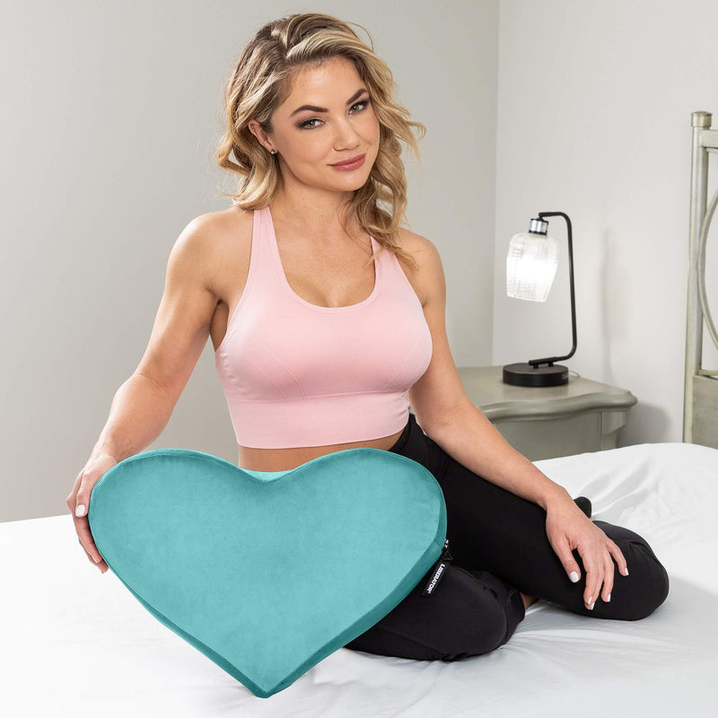 A person in a sports bra and legging is sitting on their hip on a bed. The Liberator Heart Wedge in teal is propped up against their knee to showcase its size. It is clearly wider than the person's hips and is about the same width as the person's shoulders. | Kinkly Shop