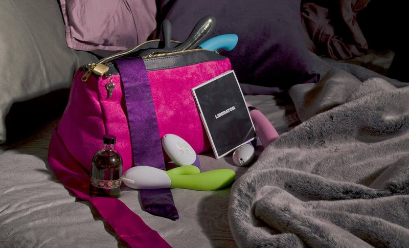 A Liberator Tallulah Locking Toy Bag sitting open on a plush bed. All of the items that were inside of the Liberator Tallulah Locking Toy Bag are tossed next to the bag. The bag is unzipped. Multiple dildos are poking out the top. Multiple toys are scattered around its bottom. A bottle of lube is sitting next to the bag as well. | Kinkly Shop
