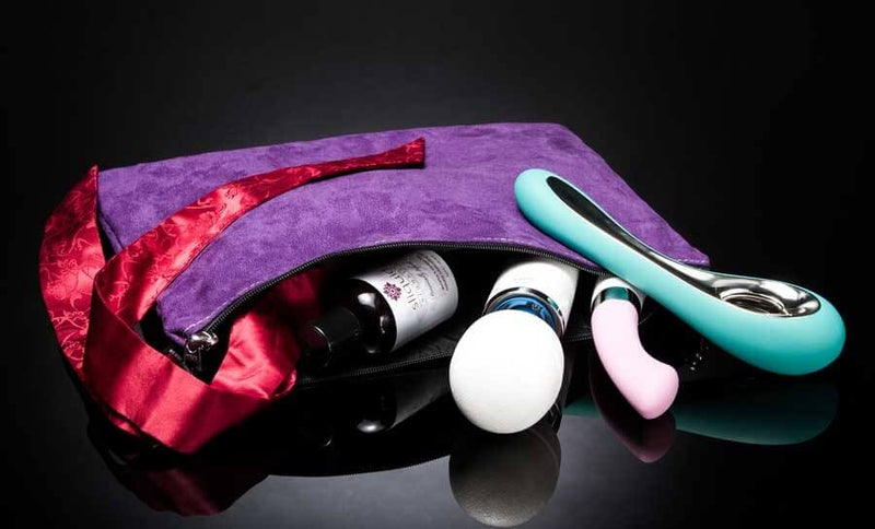 The Liberator Zappa Toy Bag in Grape is unzipped, and toys are spilling out of the open zipper. This includes a bottle of lube, a Vibratex magic wand, silky restraints, and two insertable vibrators by LELO. | Kinkly Shop
