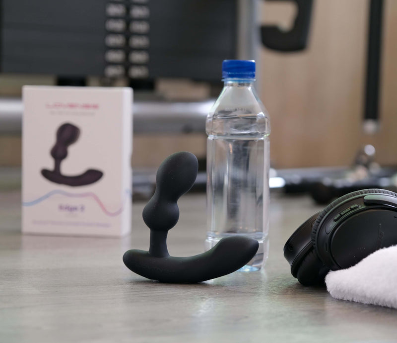 Lovense Edge 2 sits out on a desk. It's slightly taller than half the height of the water bottle it's sitting next to it, and its packaging can be seen in the background. It's also sitting up next to a set of wireless, high-end headphones and a plush, white gym towel. | Kinkly Shop