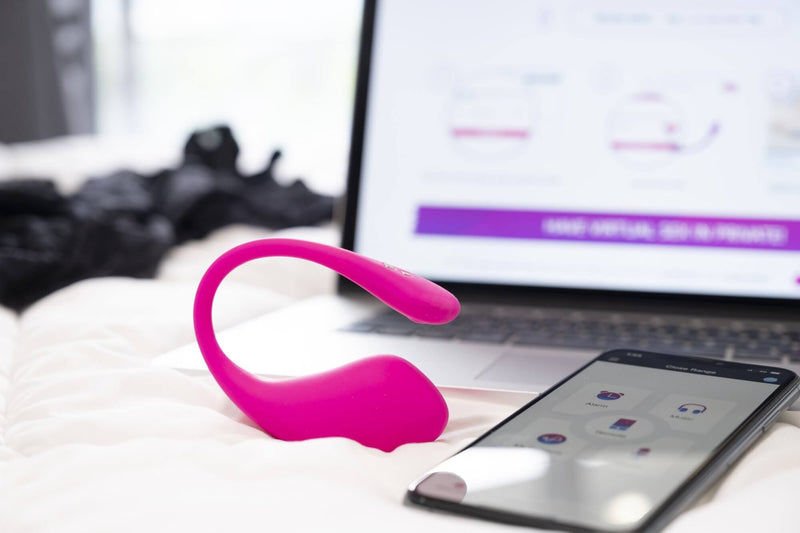 The Lovense Lush 3.0 sits next to a laptop and a cell phone in a brightly lit bedroom on a white bed. Both the laptop and the cell phone are open to Lovense apps in order to control the vibrator. | Kinkly Shop