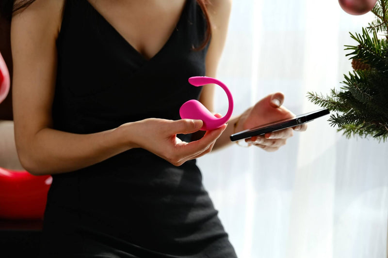 A person in a black dress is holding the Lovense Lush 3.0 in one hand and their cell phone in the other. The vibrator isn't slim, and it seems substantial compared to their hands. We assume that they're pairing the two together. | Kinkly Shop