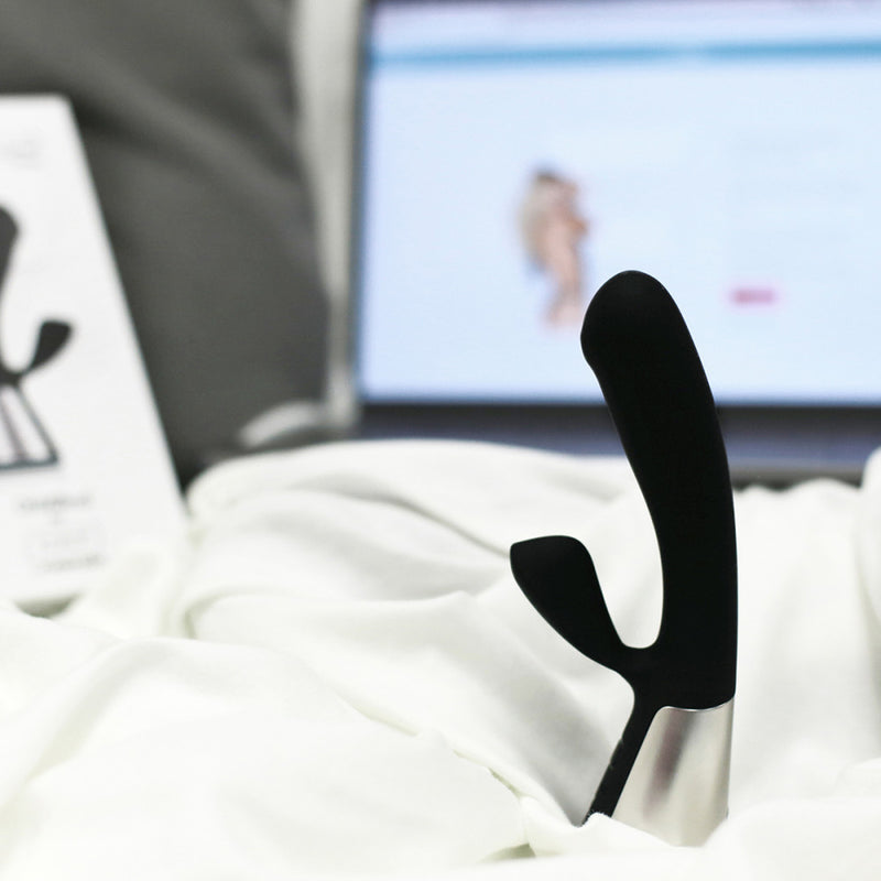 A black OhMiBod Fuse rests in white bedding while a laptop is open in the background | Kinkly Shop