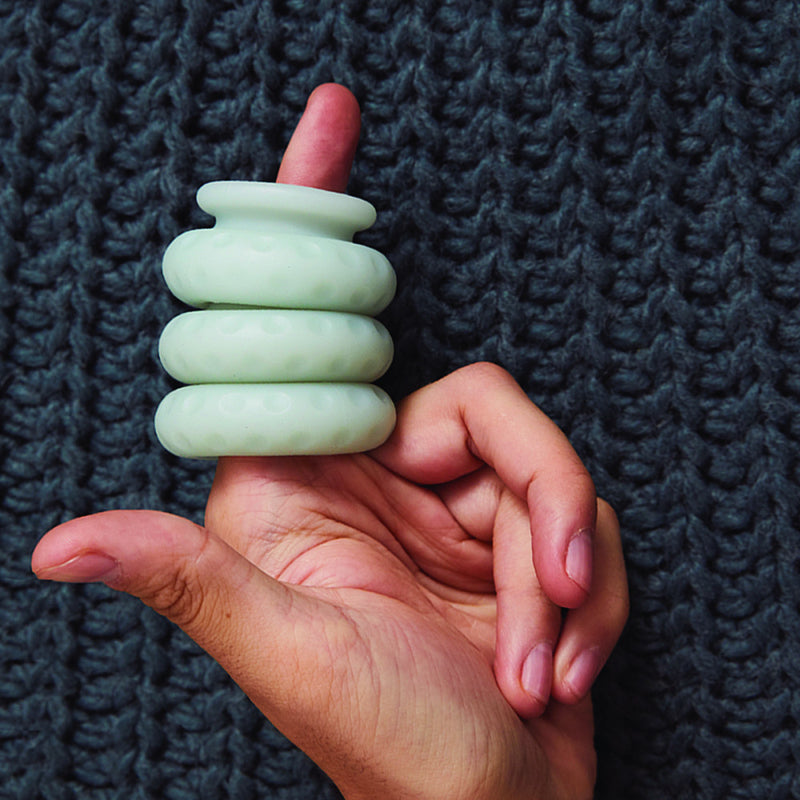 The OhNut Penetration Buffer Penis Limiter in Light Green are stacked up on a person's finger. The background is a dark blue, knitted surface that contrasts well with the light green OhNut Penetration Buffer Penis Limiter. | Kinkly Shop