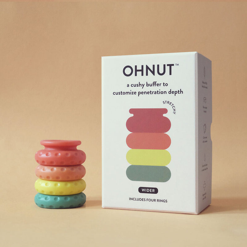The OhNut Penetration Buffer Penis Limiter in Wide Rainbow coloration sits out on a stack on a beige background. Next to it, the packaging for the OhNut Penetration Buffer Penis Limiter is sitting out. | Kinkly Shop
