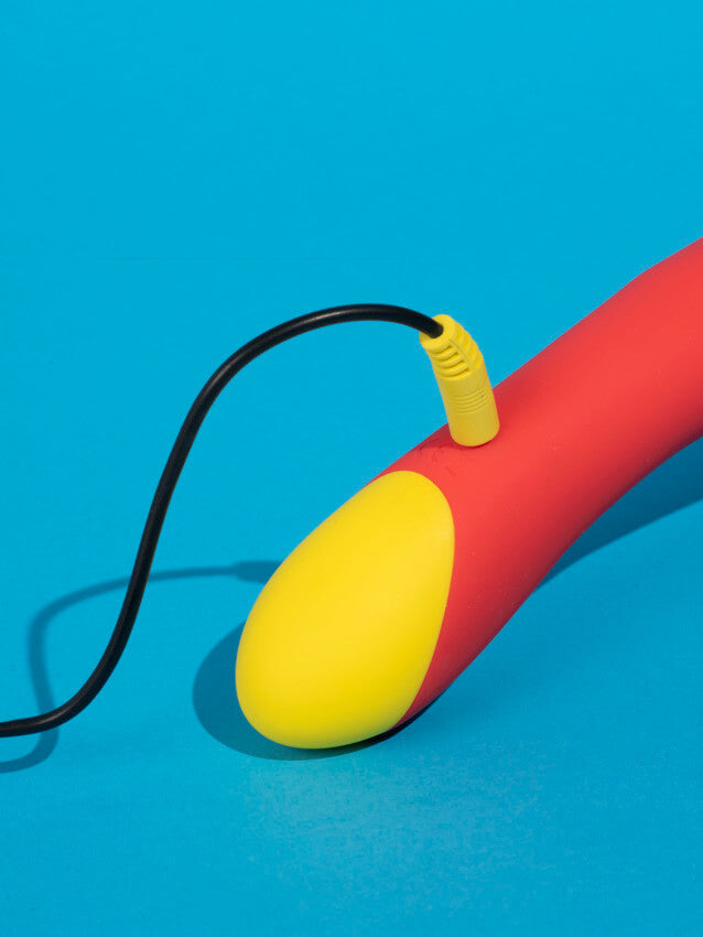 The ROMP Hype G-Spot Vibrator sitting on top of a light blue background. The color-matching USB cable is plugged into the vibrator to show how it charges. | Kinkly Shop