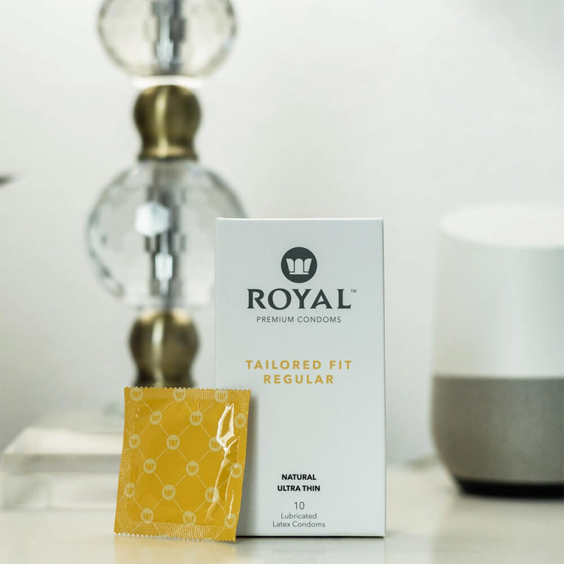 A Royal Tailored Fit Ultra Thin Vegan Latex Condoms - 10 Pack condom sits out in front of the box. Both the box and condom are sitting upright in the foreground while a glass, bulbed lamp is in the background. | Kinkly Shop