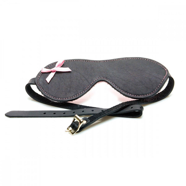 The Sex Kitten Leather Buckle Blindfold laying out on a white surface. The front of the Sex Kitten Leather Buckle Blindfold is smooth, black leather with a small, pink bow in the upper left corner of the blindfold. This image shows the buckle adjustment system for getting a good fit. | Kinkly Shop