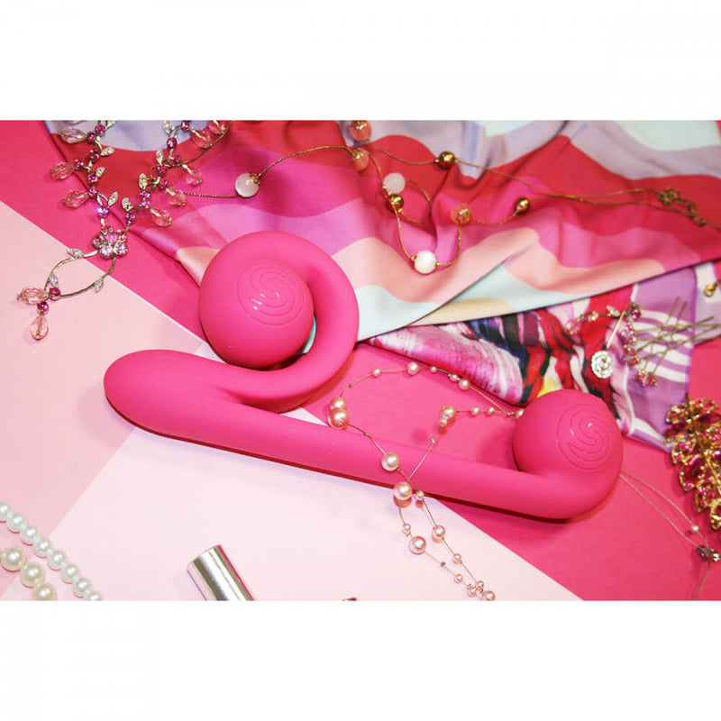 A lifestyle image of the Snail Vibe sex toy shows it laying down on top of a variety of pink, loud fabrics and jewelry. | Kinkly Shop