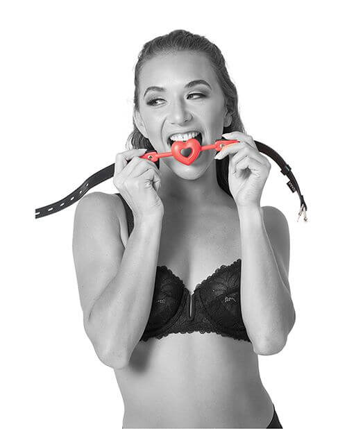 A person in lingerie is smiling as they hold the heart-shaped Sportsheets Amor Breathable Ball Gag in their mouth | Kinkly Shop