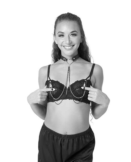 A person in lingerie is smiling at the camera while they wear the Sportsheets Sex & Mischief Amor Collar with Nipple Clamps. Their fingers are placed with their nipples would be, and the two included nipple clamps are fastened onto their fingers. | Kinkly Shop