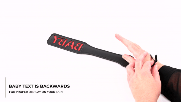 GIF showing the Sportsheets Baby Black Vinyl Paddle being handled by a person. The person picks up the paddle and slaps their hand with it. The wrist strap can be seen worn around the person's wrist. The text says "Baby text is backwards for proper display on your skin." | Kinkly Shop