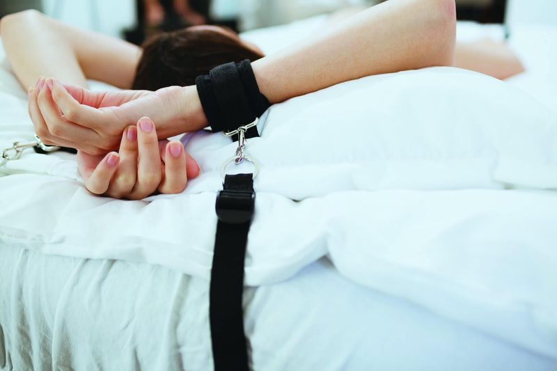 A close-up of a person's wrist wrapped in the cuff of the Sportsheets Under the Bed Restraint System. The image shows the nylon tether attached to the cuff with a clip. | Kinkly Shop