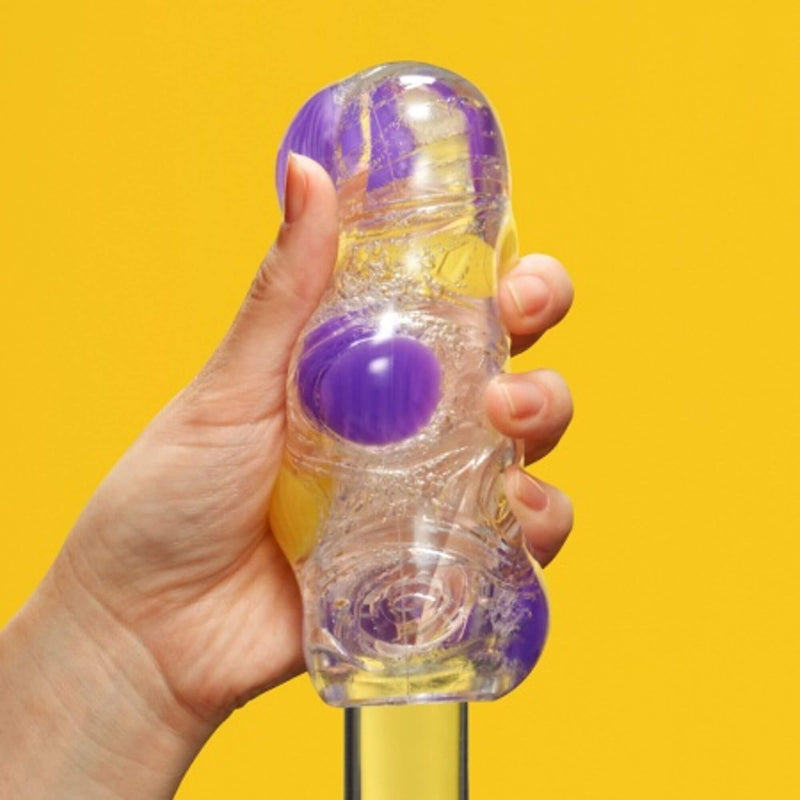 A hand holds the Tenga Bobble while the toy is being slid onto a clear, see-through glass dildo. The nodules visibly protrude away from the glass dildo to showcase the different textures. | Kinkly Shop