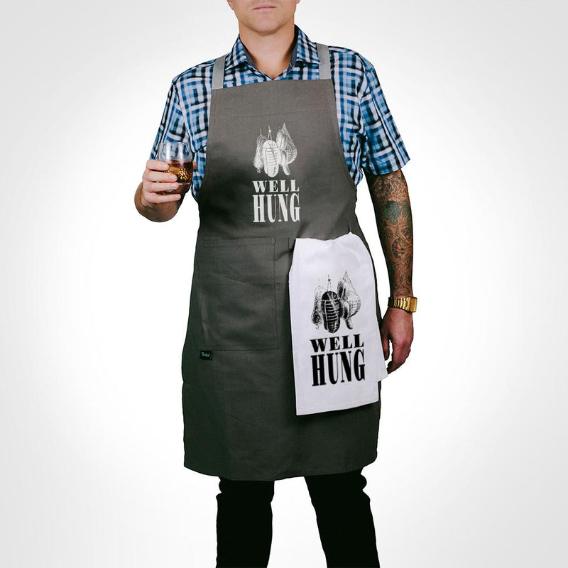 Person wearing the Twisted Wares Well-Hung Apron while holding a glass of a brown beverage with a matching towel hanging from the towel loop | Kinkly Shop