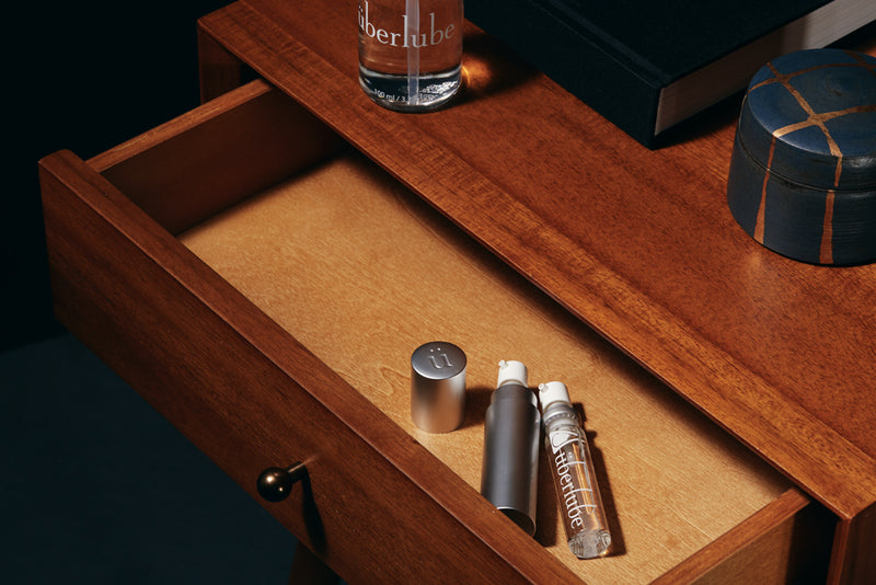 The Überlube Good-to-Go Travel Size Lube is laying inside of an empty bedside table drawer. A large Uberlube bottle size can be seen in the corner for comparison. | Kinkly Shop
