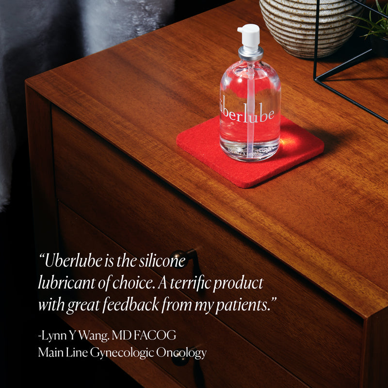 Bottle of Überlube 100ml waterproof sex lube sits out on a nightstand on top of a vibrant red coaster. The quote on the image reads "Uberlube is the silicone lubricant of choice. A terrific product with great feedback from my patients" attributed to Lynn W. Wang at the Main Line Gynecologic Oncology department. | Kinkly Shop