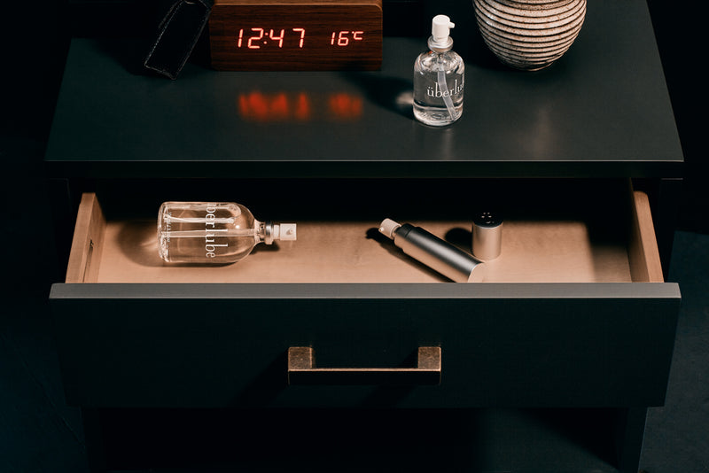 An end table is shown. The Überlube 50ml long lasting silicone lube rests on top of the end table next to a fancy wooden digital clock. Inside the open end table drawer, the 100ml Uberlube bottle is sitting next to the Good-To-Go travel size silicone lubricant Uberlube. | Kinkly Shop
