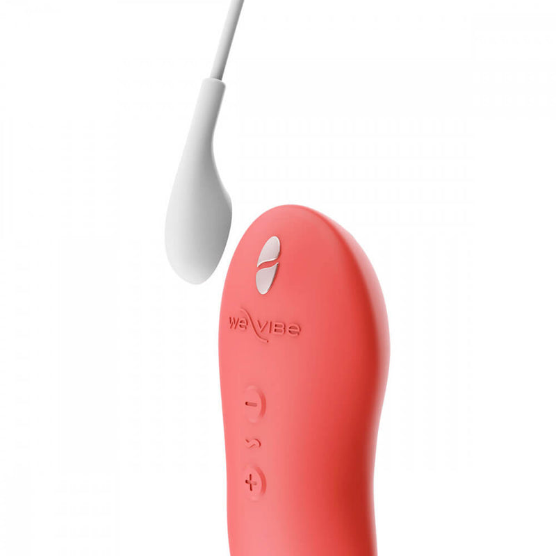 The We-Vibe Touch X in Crave Coral is laying out next to its magnetic charging cable. The cable is held slightly away from the vibrator to showcase how the two would fit together. | Kinkly Shop