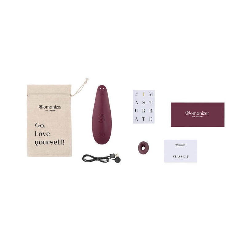 The Womanizer Classic 2 sits out with all of the items that the air suction vibrator comes with. It includes a printed drawstring bag, the charging cable, an instruction manual, a safety manual, warranty information, and an extra head in a different size to help ensure the best fit. | Kinkly Shop