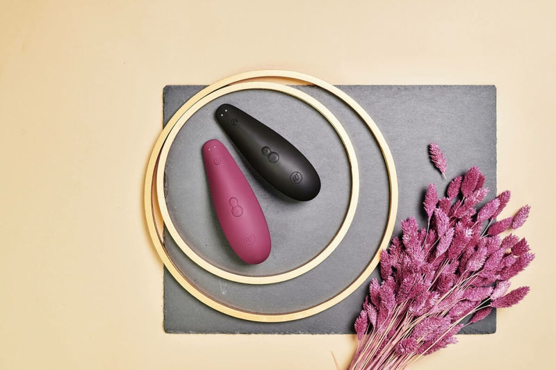 The Womanizer Classic 2 in Bordeaux and Black are both sitting next to each other inside of two gold loops. There are dried flowers placed next to them to make their colors stand out more. | Kinkly Shop