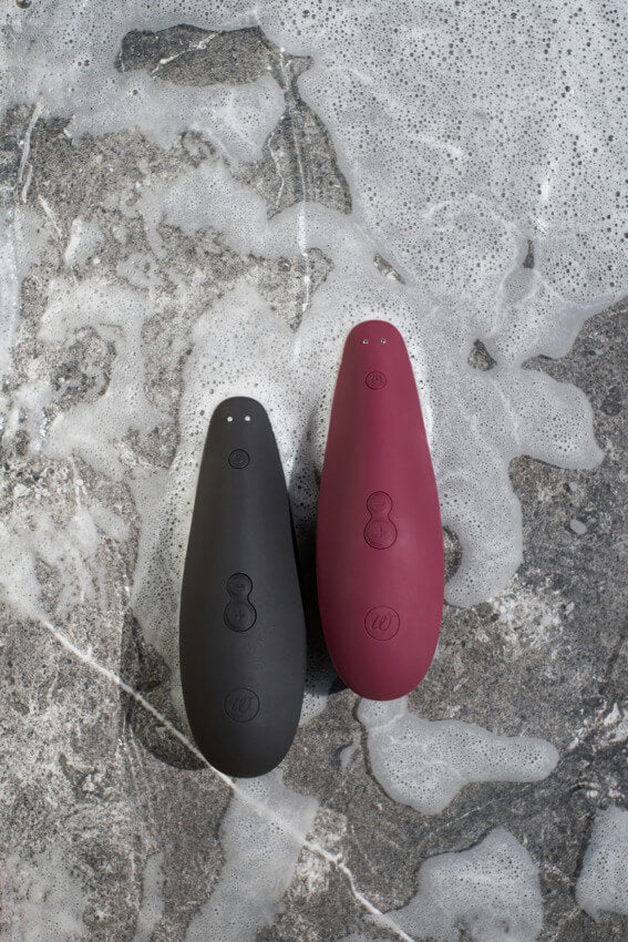 The Womanizer Classic 2 in Black and Bordeaux both sit next to each other on a concrete ground. The ground is covered in a thin layer of soapy bubbles. This demonstrate's the vibrator's waterproof capabilities and also allows for contrasting the color options. | Kinkly Shop
