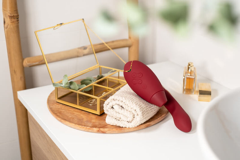The Womanizer Duo 2 in Bordeaux sits out on a sink. It sits on top of a washcloth next to a jewelry box. It a deep, vibrant red that really stands out. | Kinkly Shop